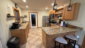 A kitchen with granite countertops, wooden cabinets, a stainless steel refrigerator, a microwave, and a stove. There are two bar stools and a trash can. - Mountain Lake House - Hot tub, fire pit, fish and swim! (Almond)