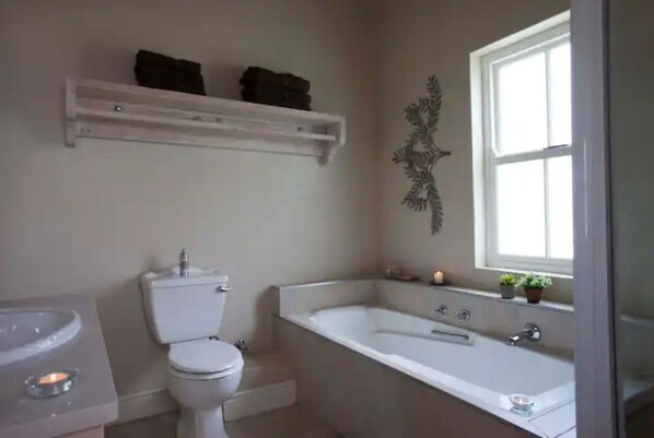 A bathroom with a toilet, sink, and bathtub. There is a shelf above the toilet and a decorative wall piece above the bathtub. - Luxurious Country Lodge. (Stanford)