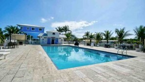A swimming pool with a tiled deck, a blue building in the background, and palm trees. - Key Largo 2 bedroom/1 bath with community pool (Key Largo)