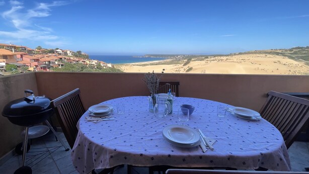Une table ronde recouverte d’une nappe à motifs floraux, mise avec des assiettes, des couverts et un pichet, d’où l’on peut admirer un paysage côtier avec des bâtiments et des dunes de sable.