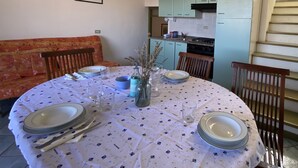 Un coin repas avec une table ronde dressée pour quatre personnes, entourée de chaises en bois. On aperçoit un canapé au fond et une cuisine à travers une porte ouverte.
