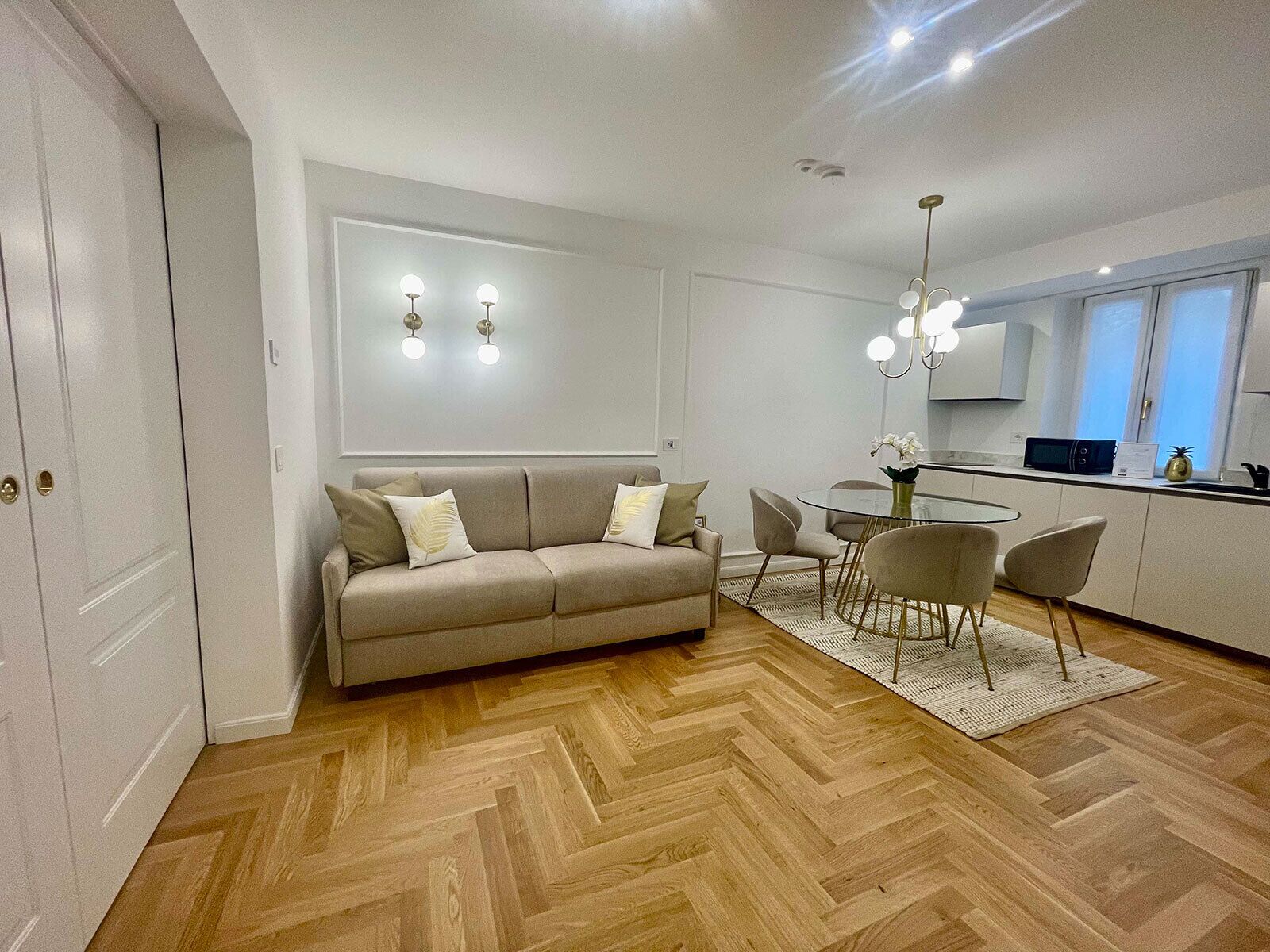A modern living room with a sofa, a dining table, and chairs. There is a kitchen area with a microwave and a refrigerator.