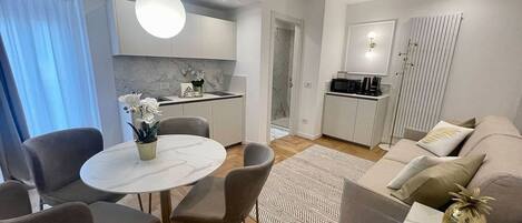 Una sala de estar moderna con una mesa de comedor redonda, sillas grises, un sofá con cojines, una máquina de café y una zona de cocina con un microondas.