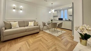 A modern living room with a sofa, a dining table, and chairs. There is a kitchen area in the background with a microwave and a refrigerator. - Bellagio Luxury Suites Apartments (Bellagio)