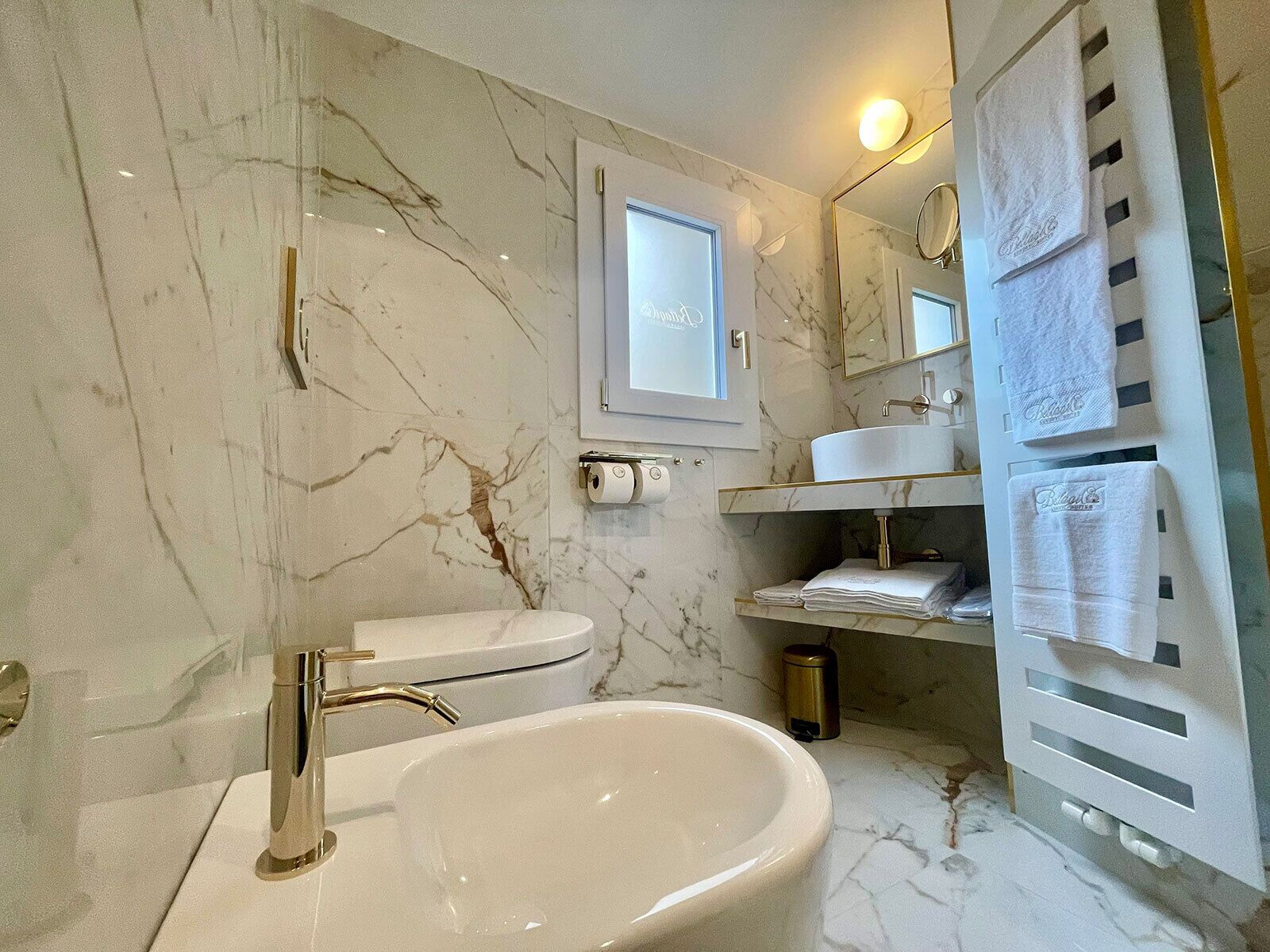 A modern bathroom with a marble wall, a white sink, a gold faucet, a toilet, and a towel rack.