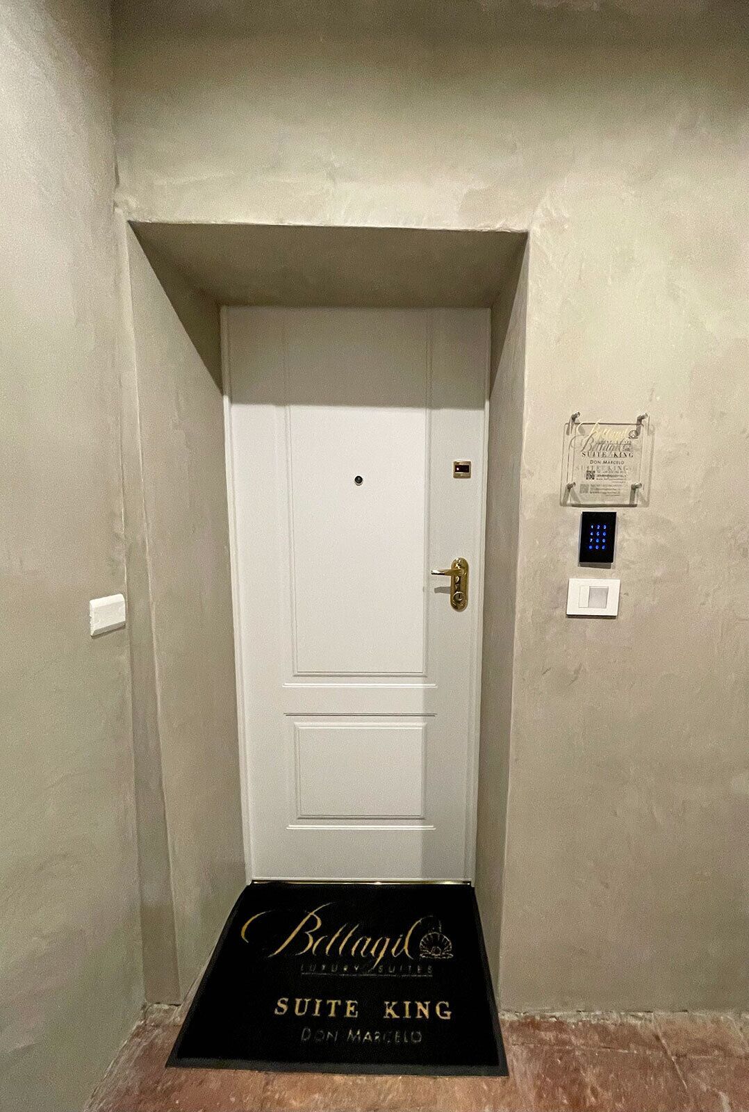 A white door with a digital lock and a keyhole, a doormat with "Bottagel Suite King Don Marcelo" text, and a small plaque on the wall.