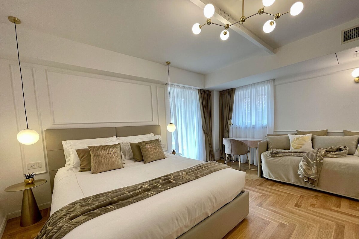 A modern hotel room with a large bed, a sofa, a desk, and a chandelier.