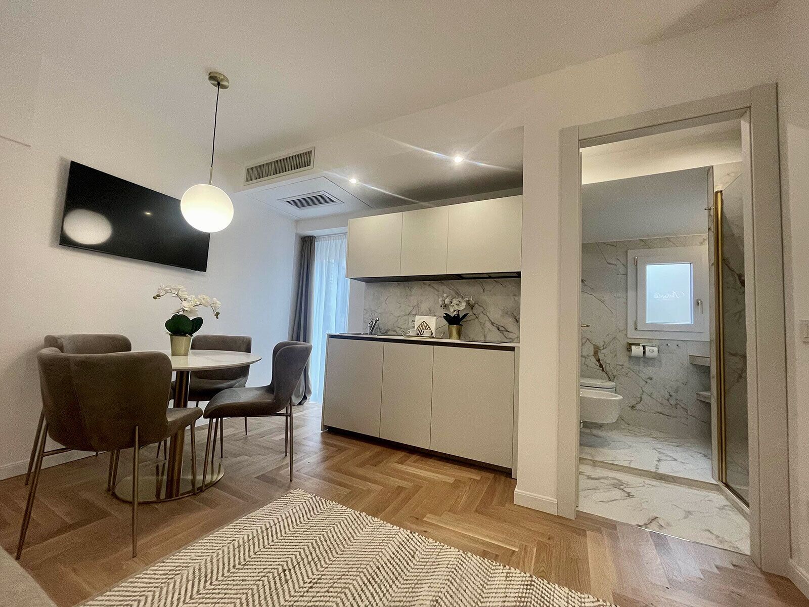 A modern kitchen with white cabinets, a marble countertop, and a built-in oven. There is a dining area with a table and chairs, and a large window providing natural light.