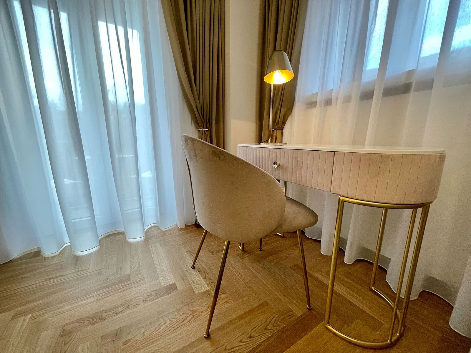 Ein Zimmer mit Holzboden, einem beigen Sessel, einem Schreibtisch mit Lampe und durchsichtigen Vorhängen.