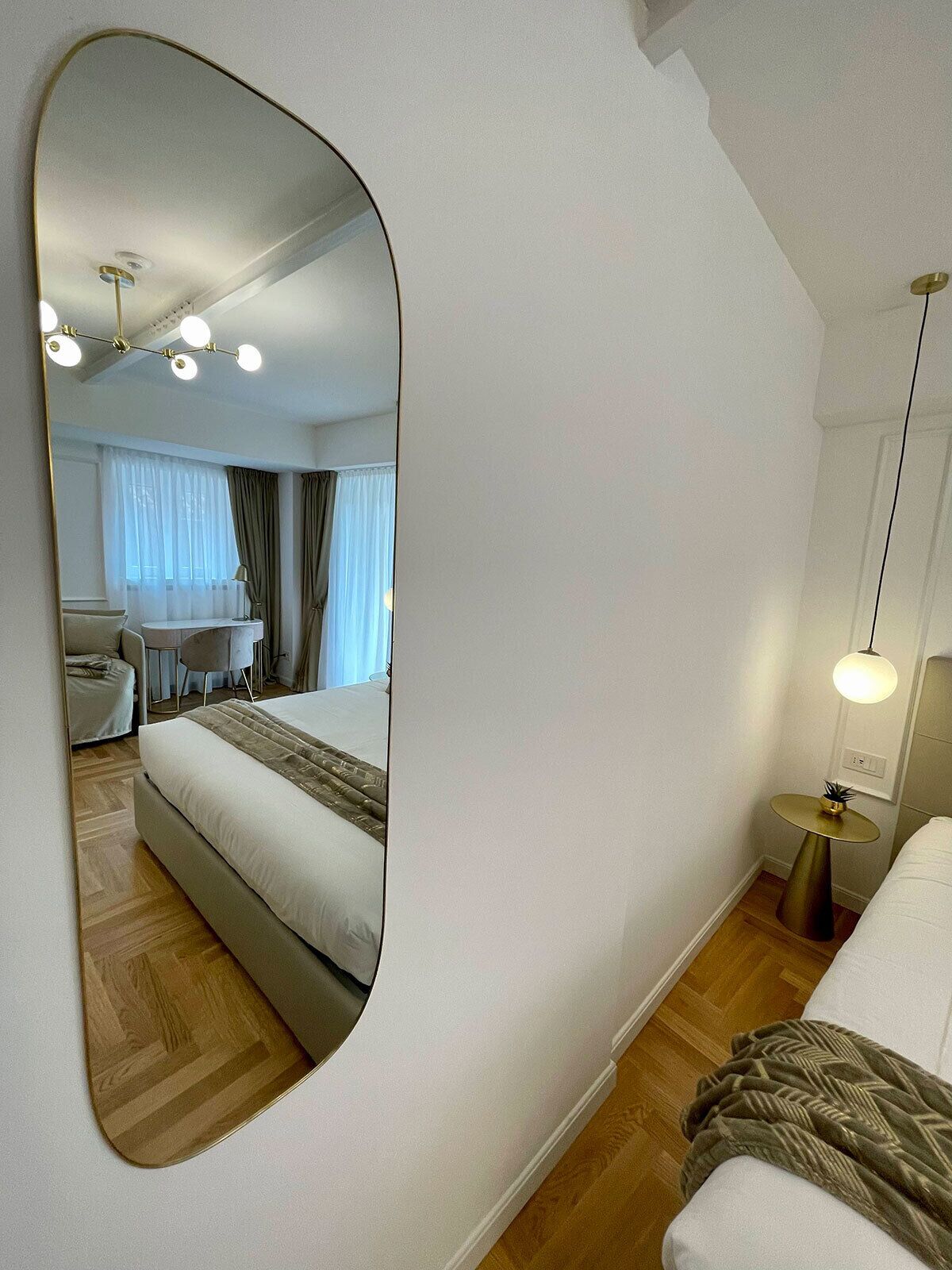 A modern hotel room with a large mirror, a bed, a bedside table with a lamp, a small round table with a plant, and a ceiling light fixture.