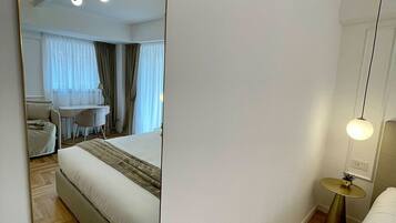 A modern hotel room with a large mirror, a bed, a bedside table with a lamp, a small round table with a plant, and a ceiling light fixture.