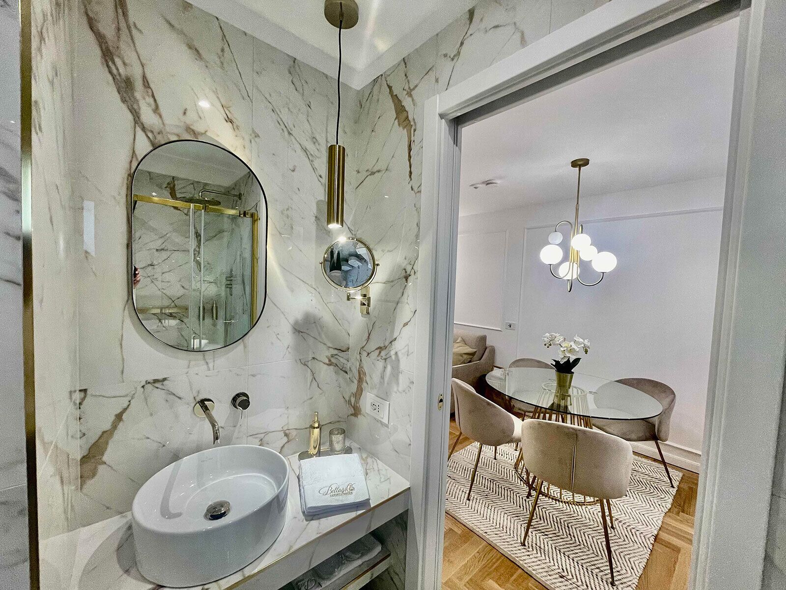 Un baño moderno con lavamanos de mármol, un espejo grande y un espejo redondo. Hay un tocador con una toalla y un pequeño jarrón de flores.