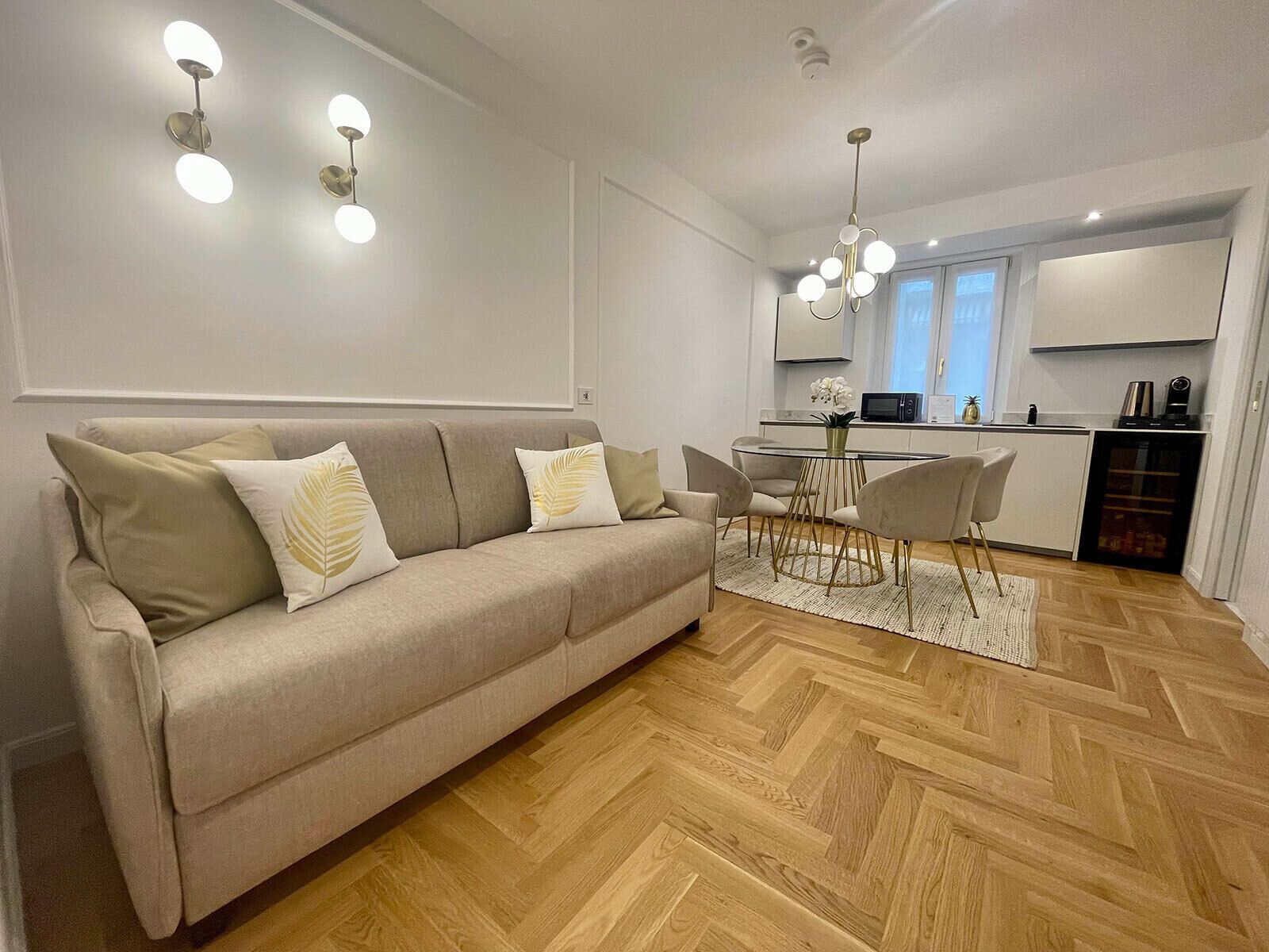 A modern living room with a beige sofa, a glass dining table, and a kitchen area with a microwave and a coffee machine.