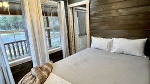 A wooden cabin bedroom with a bed, curtains, and a window overlooking a deck. - Pin Oak at Forest Villas: Swing Fire Pit Cabin, Pet-Friendly,  Cook Forest (Marienville)