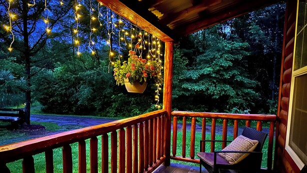 A wooden deck with a railing, a hanging flower pot, and a chair with a cushion. - Pin Oak at Forest Villas: Swing Fire Pit Cabin, Pet-Friendly,  Cook Forest (Marienville)