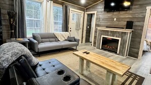 Ein modernes Wohnzimmer mit grauem Sofa, einem schwarzen Ledersessel, einem Holztisch, einem Kamin und einem an der Wand montierten Fernseher.