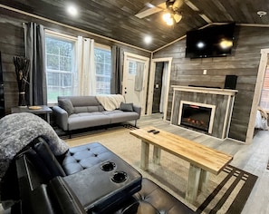 A modern living room with a grey sofa, a black leather chair, a wooden coffee table, a fireplace, and a TV mounted on the wall.