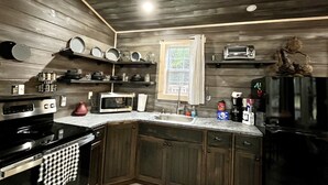 A kitchen with wooden walls, a black stove, a microwave, a sink, and various kitchen items on shelves. - Pin Oak at Forest Villas: Swing Fire Pit Cabin, Pet-Friendly,  Cook Forest (Marienville)