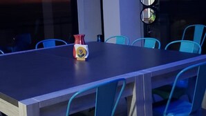 Moderni ruokailutila, jossa on pitkä pöytä ja siniset tuolit, koristeellinen maljakko sekä suuri ikkuna, josta avautuu näkymä auringonlaskuun.