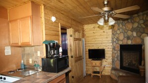 Interno di una baita in legno con cucina, dotata di piano cottura, lavello e macchina da caffè. È presente un camino in pietra e un televisore fissato al muro.