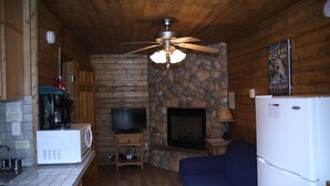 A cozy cabin interior with a stone wall, a fireplace, a television, a ceiling fan, a refrigerator, and a blue couch. - Located on our Private Fishing Pond (Red River)