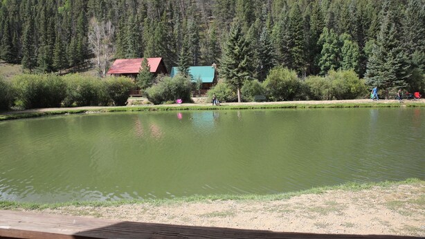 Un lac avec une terrasse en bois au premier plan, entouré d’une forêt dense et de deux bâtiments aux toits rouges.