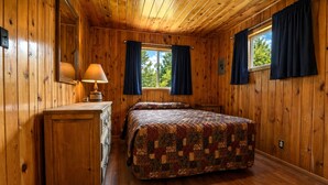 Dormitorio de cabaña de madera con cama, cómoda y ventanas con cortinas.