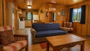 Interior de una cabaña de madera con sofá, sillón, mesa de comedor, zona de cocina y vistas al exterior.