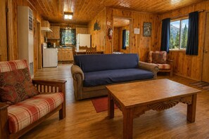 L'intérieur d'un chalet en bois comprend un canapé, un fauteuil, une table à manger, un coin cuisine et offre une vue sur l'extérieur.