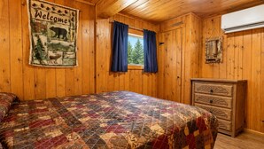 Ein Zimmer in einer Holzhütte mit Bett, Kommode, einer Willkommensschild und einem Fenster mit Vorhängen.
