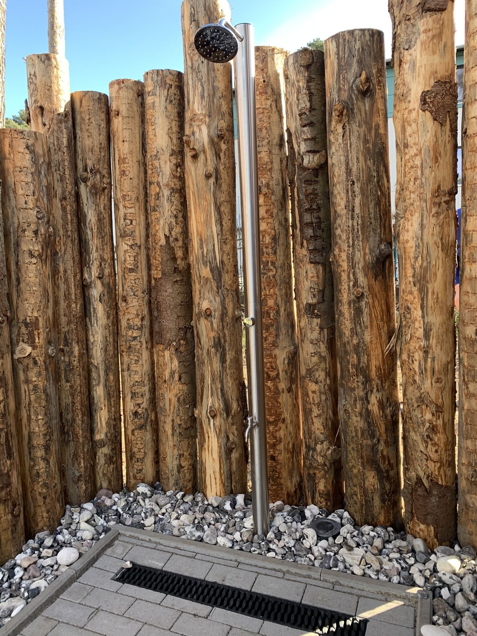 A metal showerhead mounted on a pole, surrounded by a wooden fence and pebbles.