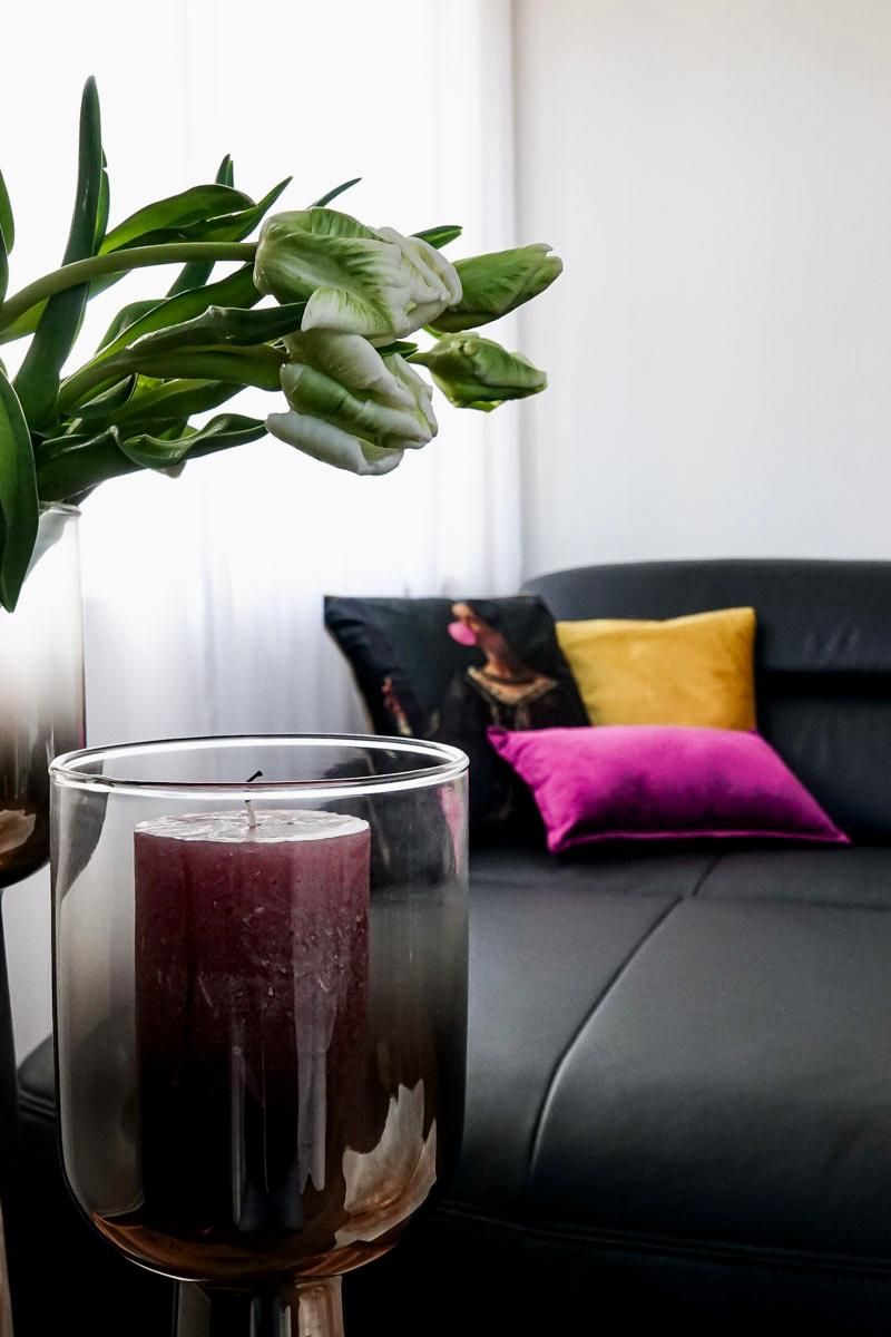 Ein Glaslaternenhalter mit Kerze, ein Topfpflanze mit grünen Blättern und eine schwarze Couch mit bunten Kissen im Hintergrund.