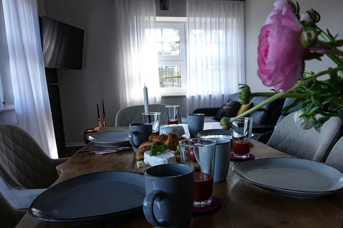 Ein Esstisch ist gedeckt mit Tellern, Tassen und Gläsern, mit einem Blumenstrauß und einer Kerze darauf.