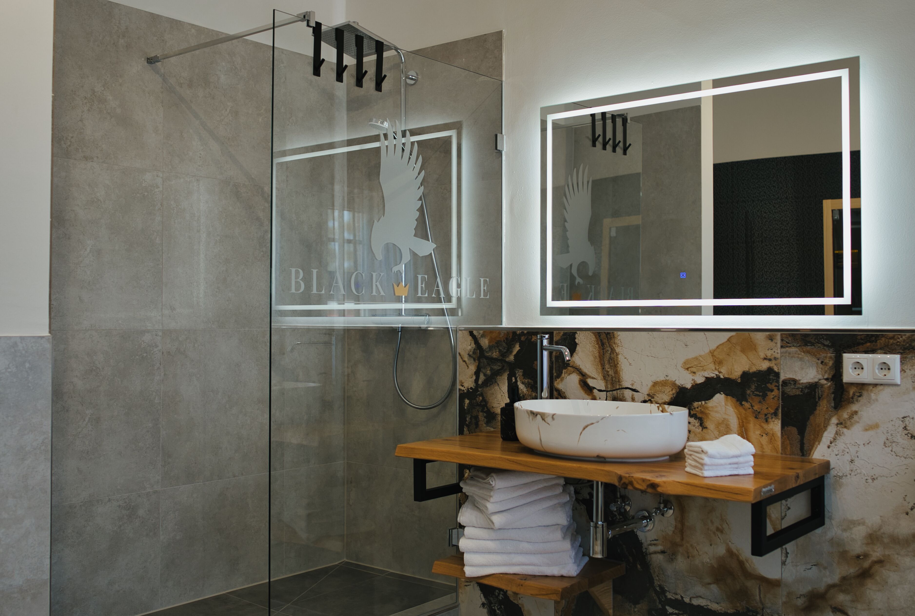 Un baño moderno con cabina de ducha de vidrio, un lavamanos blanco con salpicadero de mármol y un estante de madera con toallas dobladas.
