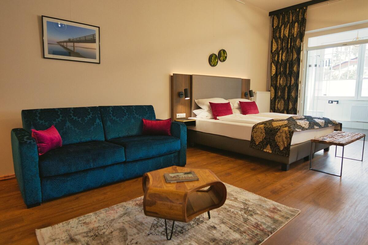 A hotel room with a blue sofa, a wooden side table, a bed with a patterned bedspread, and a view of a bridge through the window.