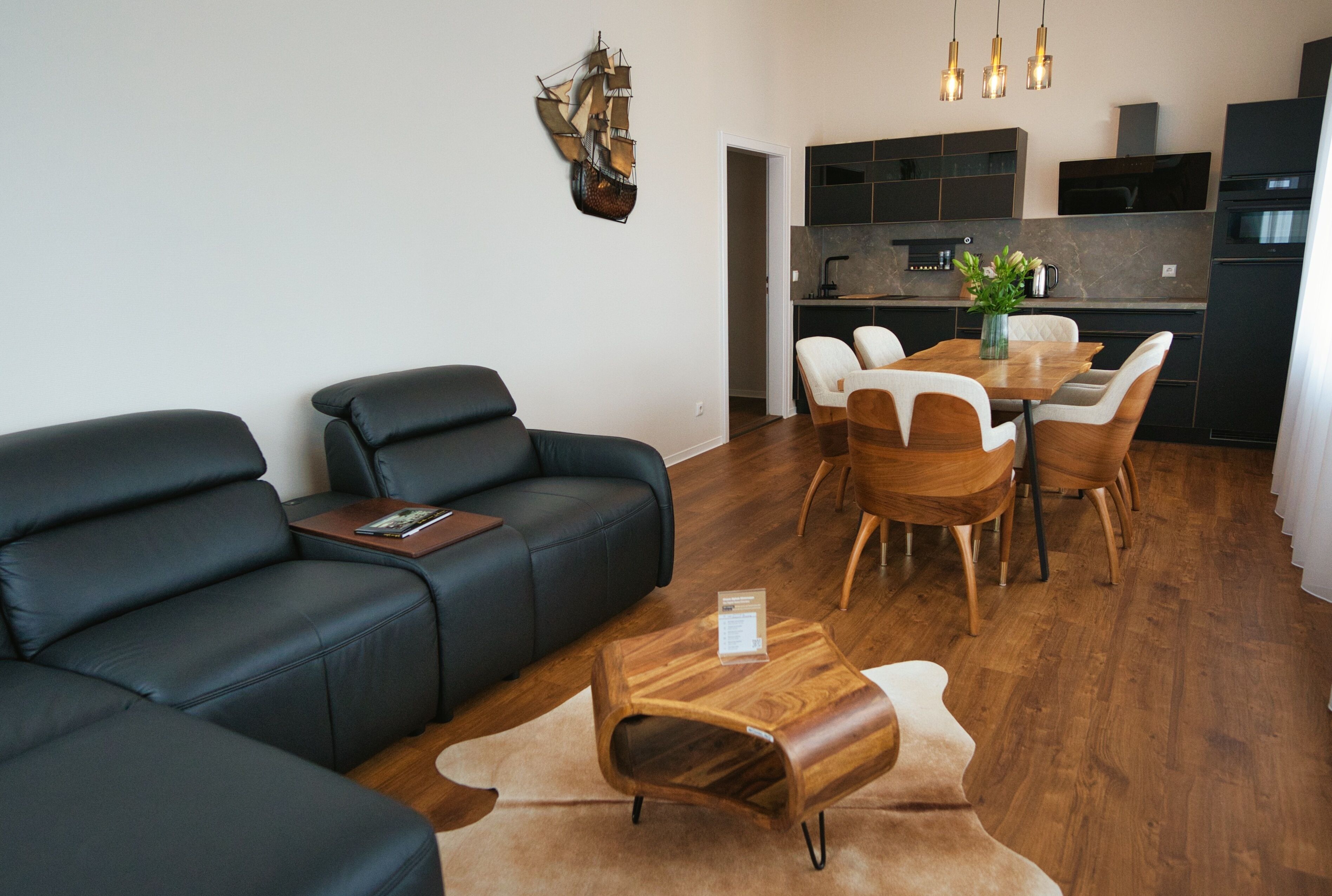 A modern living room with a black leather sofa, wooden dining table, and chairs. A kitchen is visible in the background.