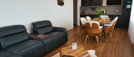 A modern living room with a black leather sofa, wooden dining table, and chairs. A kitchen is visible in the background.