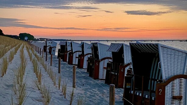Un hilero de cabañas de playa con números en las puertas, una playa arenosa y una puesta de sol sobre el agua.