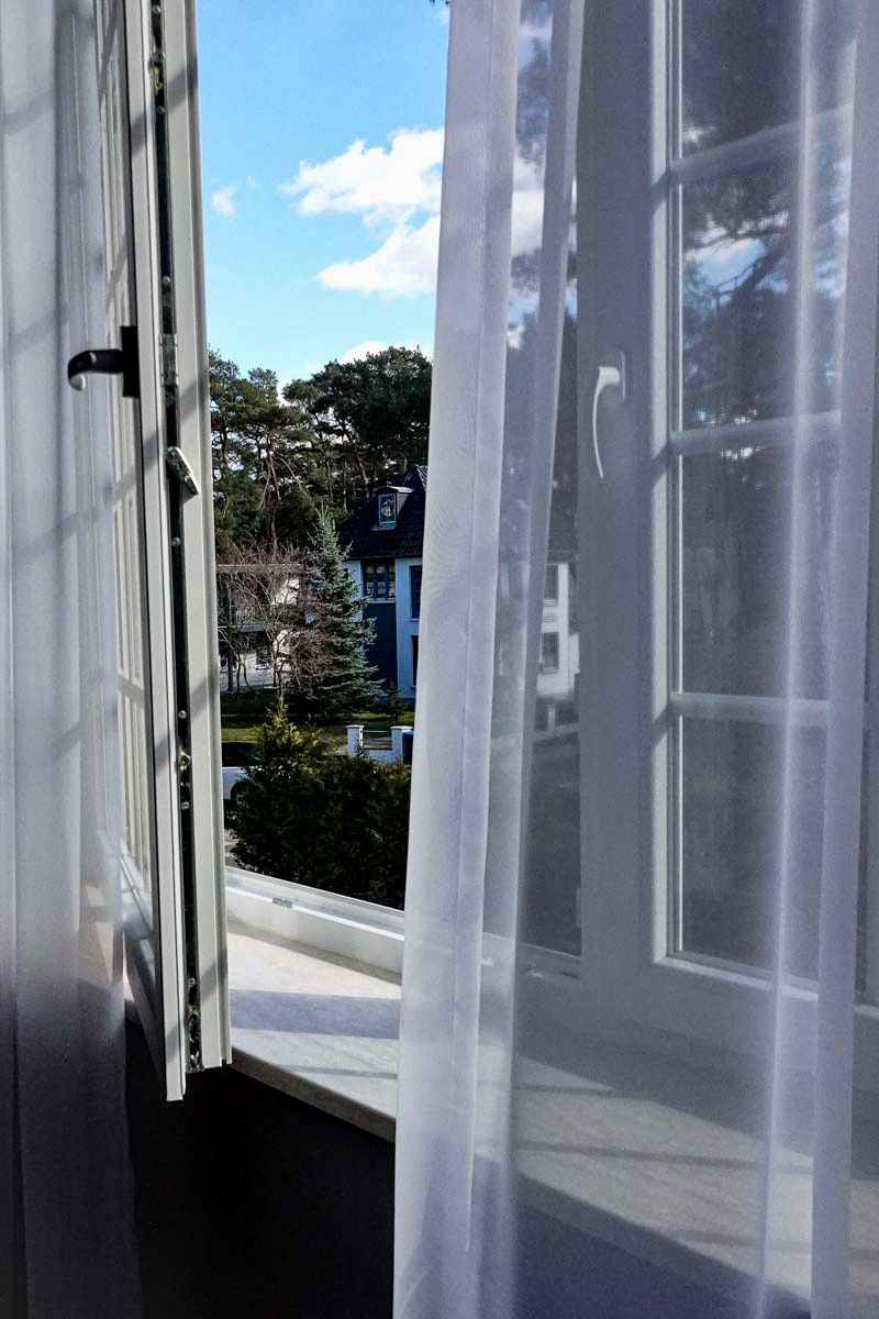 Ein Fenster mit transparenten Vorhängen, das einen Blick auf eine Wohngegend mit Häusern und Bäumen freigibt.
