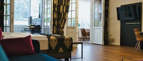 Un salon moderne avec un canapé, une table basse et un téléviseur. Il y a une grande fenêtre avec des rideaux, une porte donnant sur une autre pièce et une petite table avec une lampe.