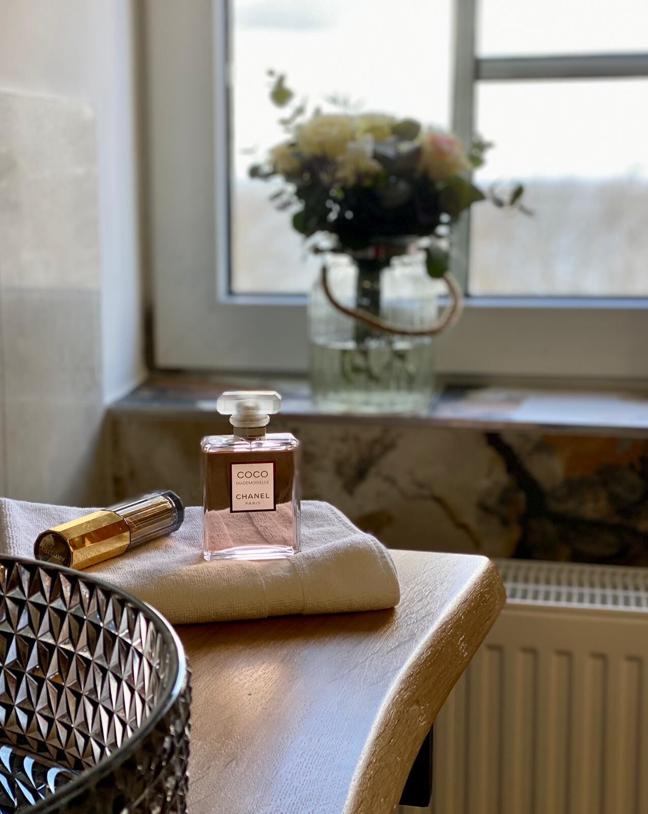 A bottle of Coco Chanel perfume on a beige cushion by a window with a vase of flowers.
