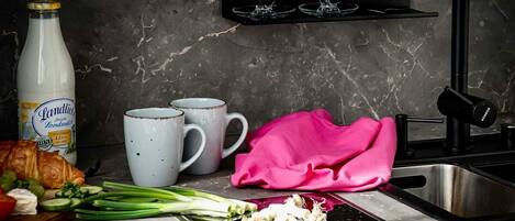 A kitchen counter with a bottle of milk, two mugs, a pink cloth, a knife, and some vegetables.