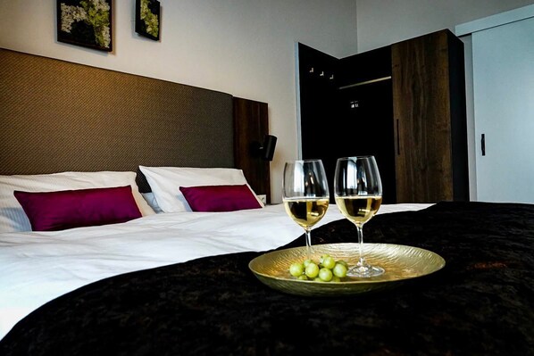 A neatly made bed with white linens and two purple pillows, a bedside table with two glasses of white wine and a plate of green grapes, and a dark headboard. - Residence Black Eagle Lubmin (Lubmin)