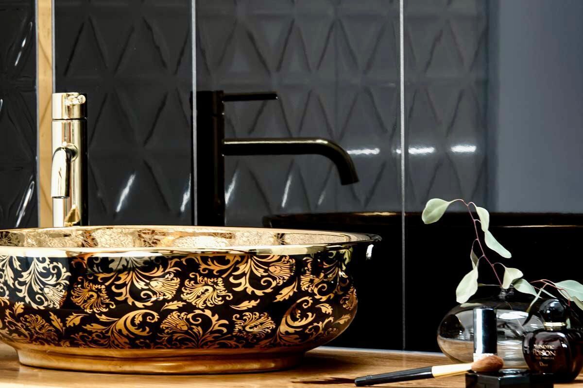 A black and gold ornate bowl on a wooden surface, a black faucet, and a glass vase with a plant.