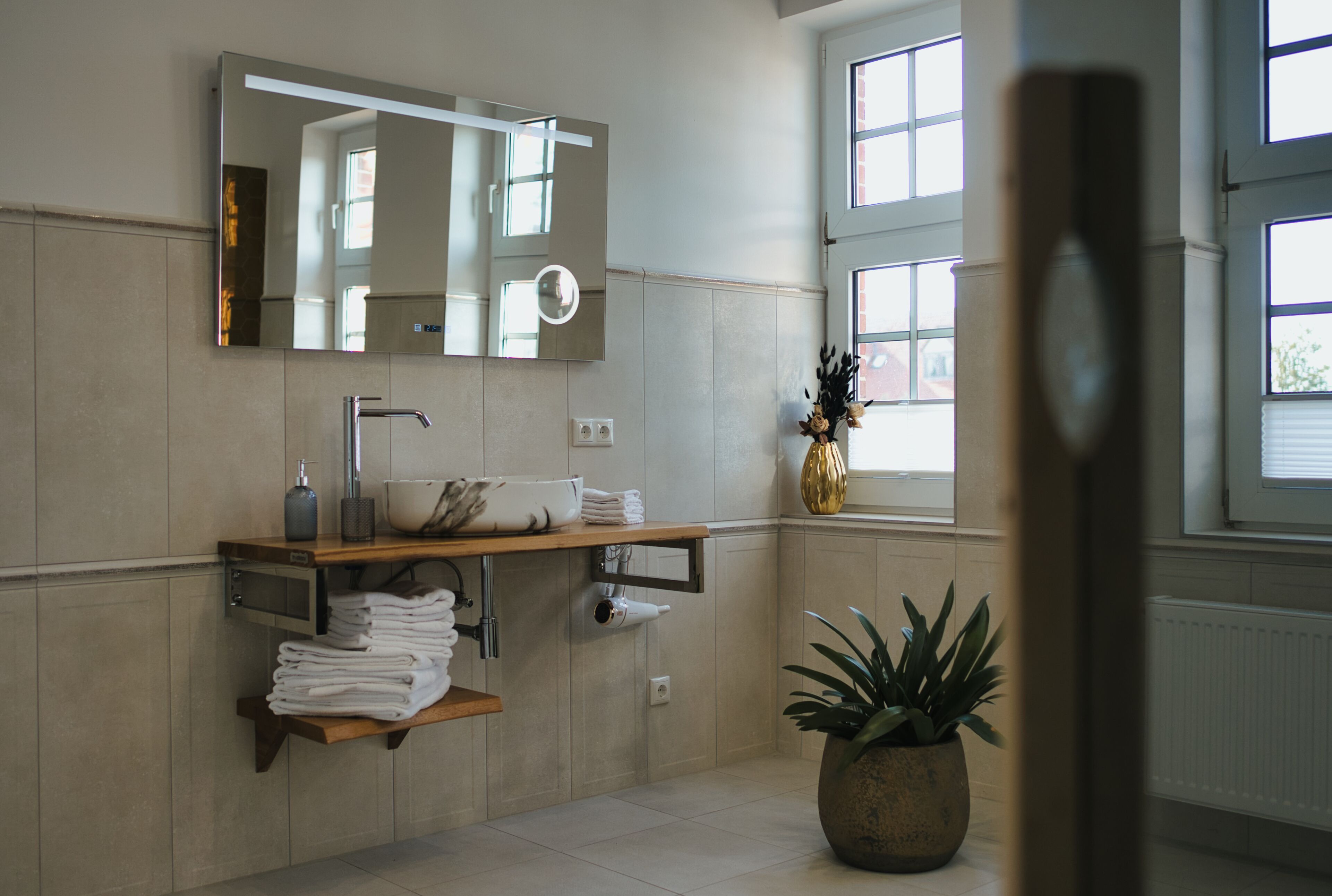 A modern bathroom with a large mirror, a sink, a towel rack, and a potted plant.