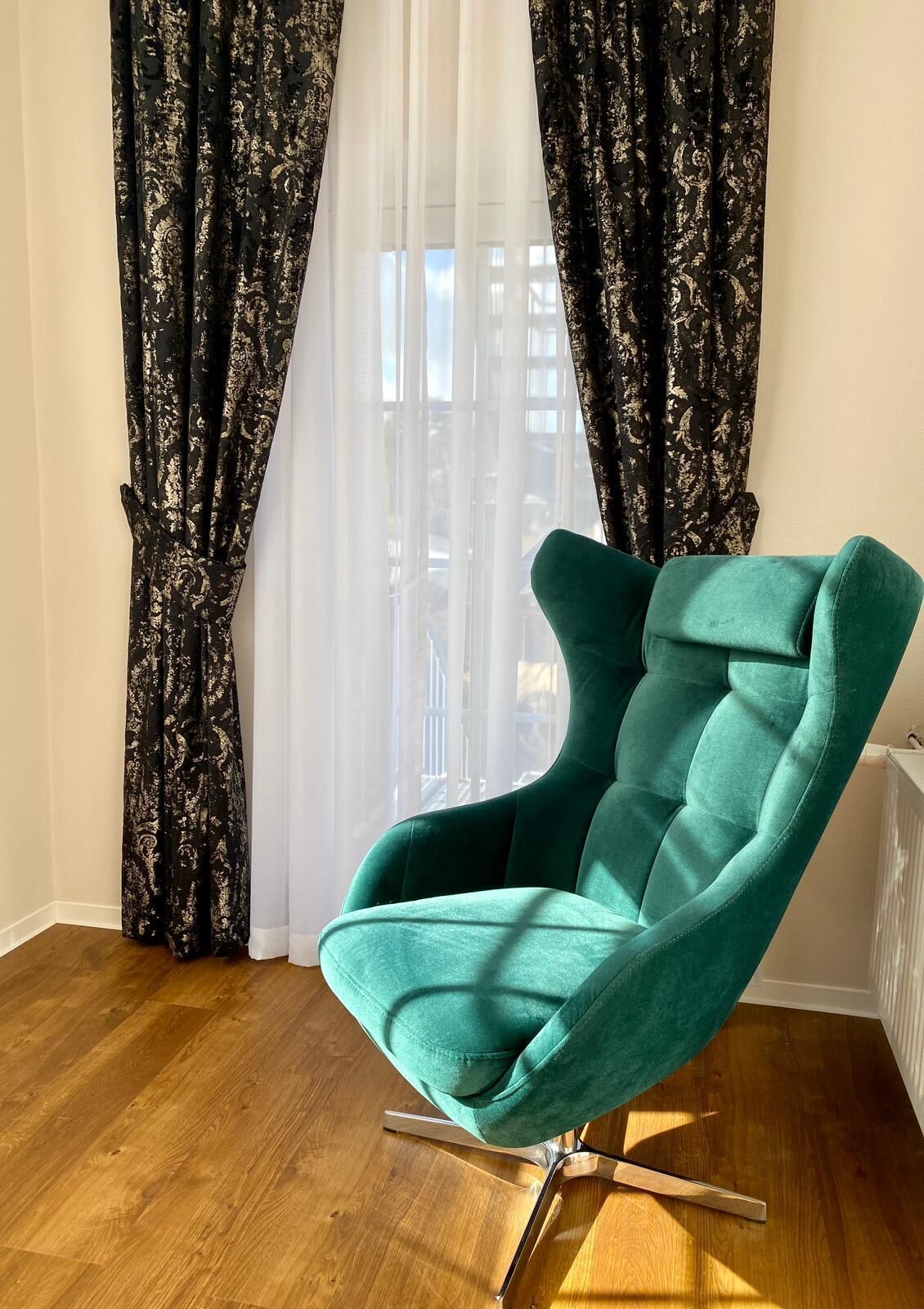 Un fauteuil de couleur turquoise avec un pied en métal, placé devant une fenêtre ornée de rideaux blancs transparents et de rideaux noirs à motifs.