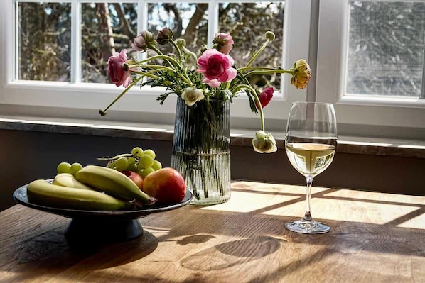Una mesa con un frutero, un jarrón con flores y una copa de vino cerca de una ventana.