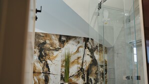 Un baño moderno con pared de mármol, cabina de ducha de vidrio y una planta en una maceta geométrica.