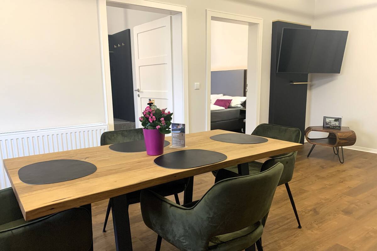 A dining area with a wooden table, black placemats, green upholstered chairs, a potted plant, and a small table with a picture frame.