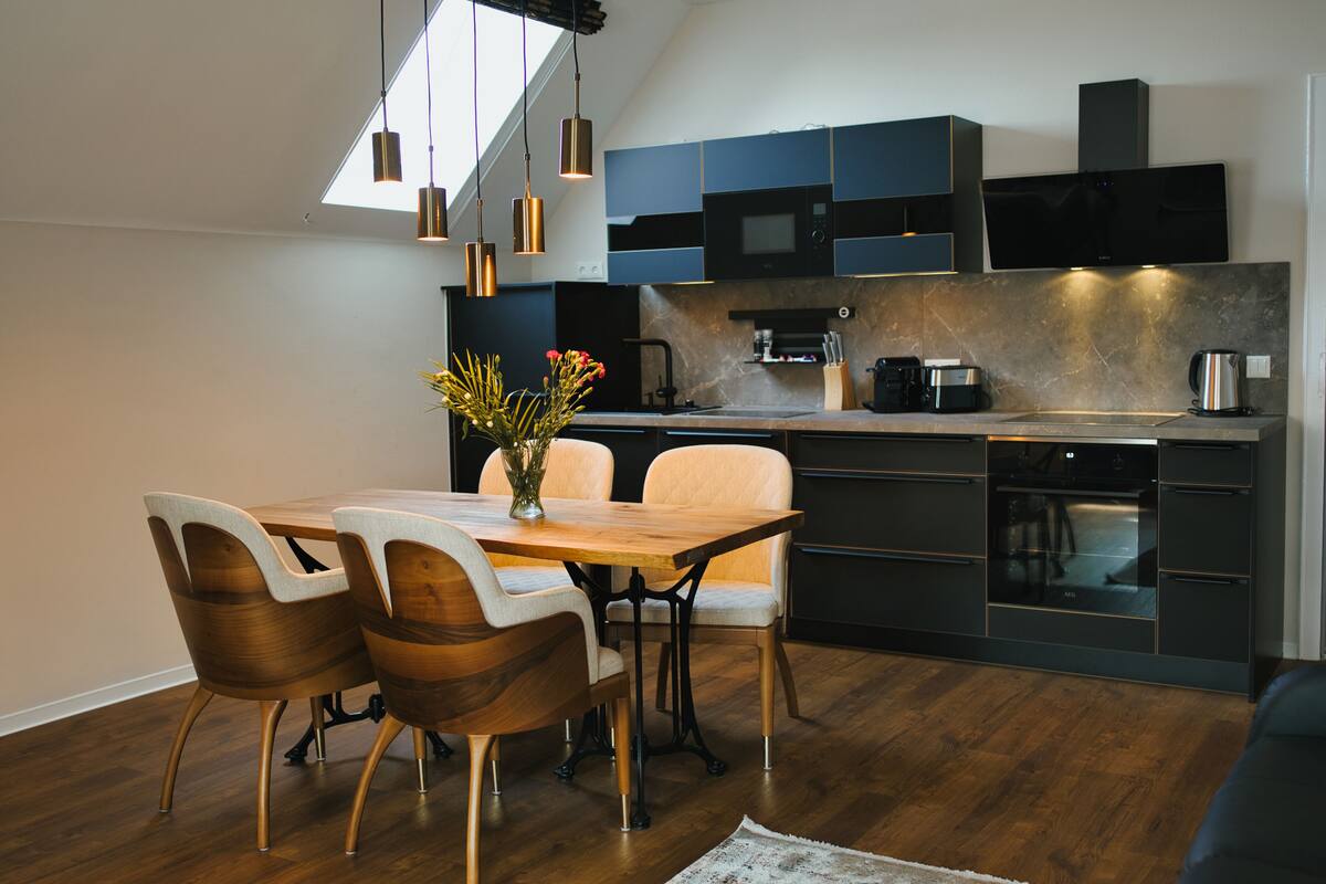 A modern kitchen with dark cabinetry, a wooden dining table, and pendant lighting.